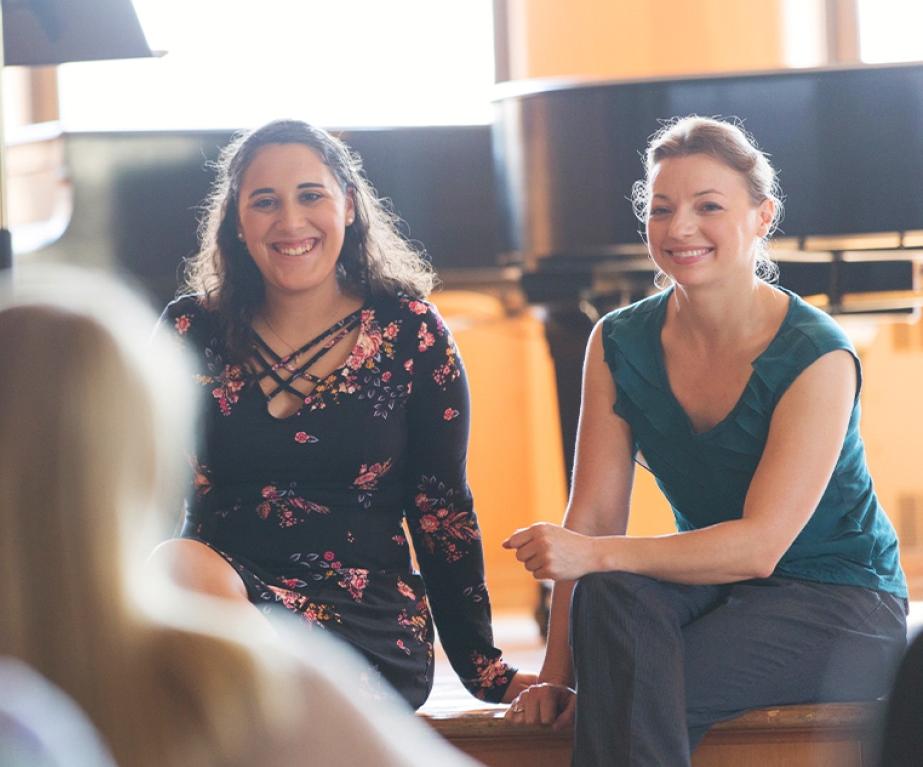 A music teacher and student sit on a stage in front of a grand piano while smiling to other students in the audience.